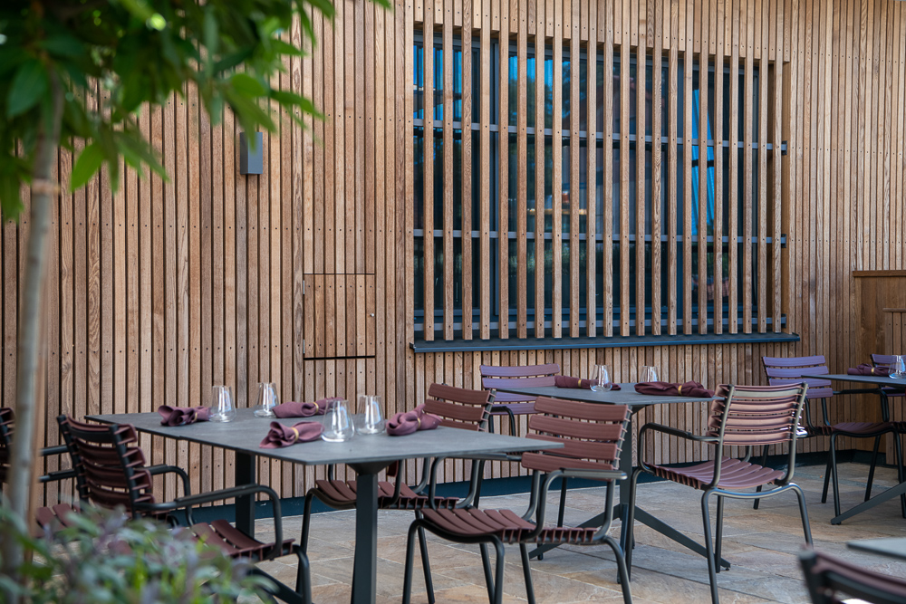 Nahaufnahme der Terrasse im Hotel Restaurant zum Oxn mit vertikaler Holzfassade in Gardasee-Ausführung von Mareiner. Gedeckte Tische mit modernen Stühlen stehen auf Steinboden vor einer strukturierten Lamellenfensterfront aus Holz.