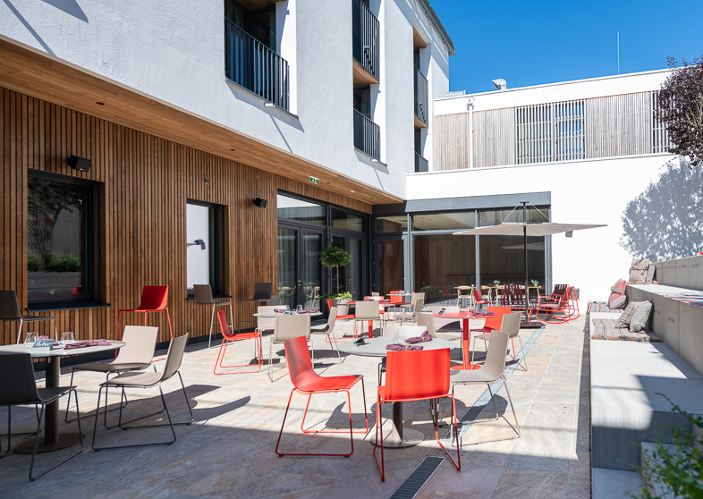 Moderne Hotelterrasse des „Hotel Restaurant zum Oxn“ mit vertikaler Holzfassade in Gardasee-Ausführung von Mareiner. Farbige Stühle und Tische auf heller Steinplatte schaffen eine lebendige Atmosphäre im sonnigen Innenhof mit gemütlichen Lounge-Ecken.