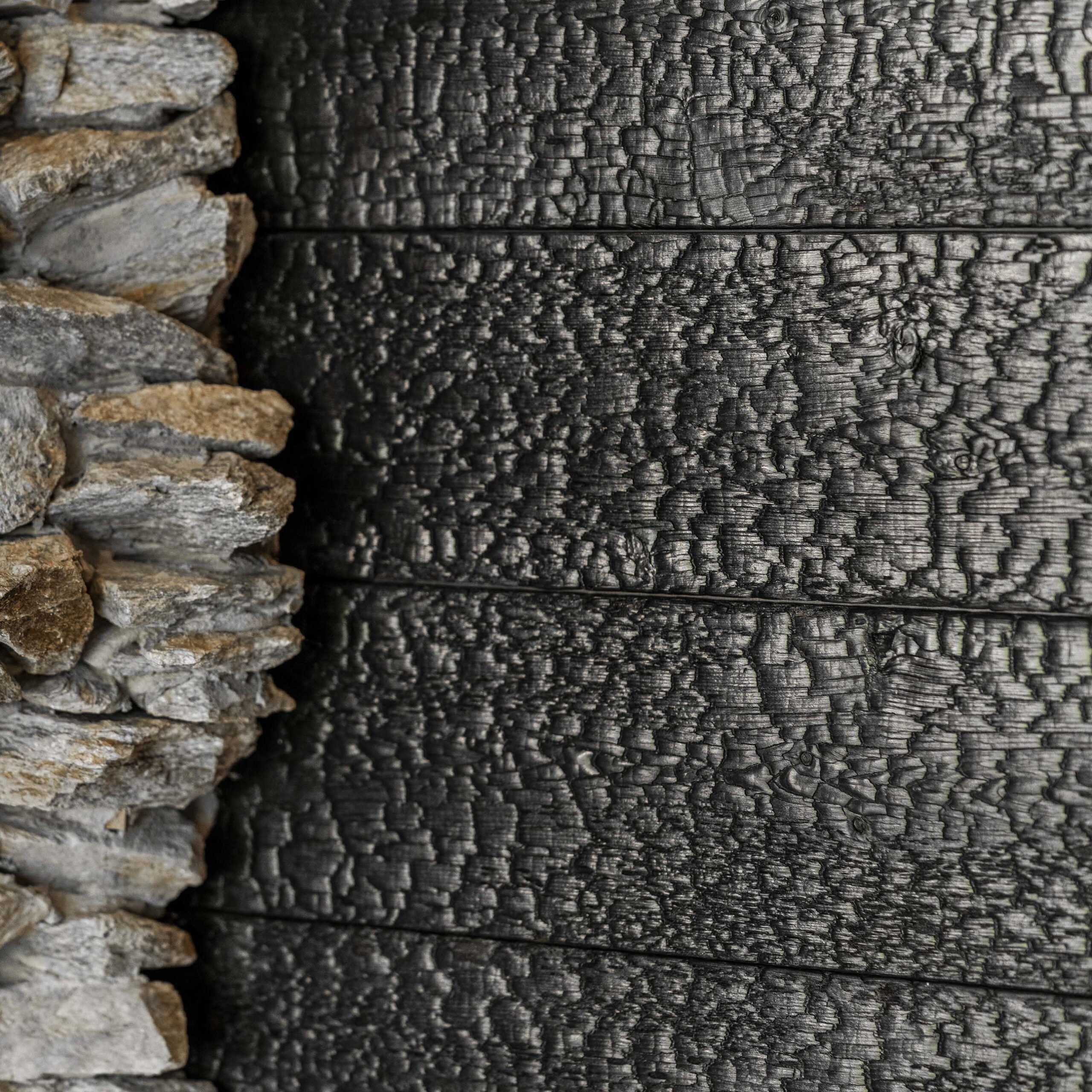 Detailansicht einer verkohlten Holzfassade aus Mareiner Vulcano beim Hotel Restaurant Krainer in Langenwang. Die markante Schuppenstruktur kontrastiert mit einer angrenzenden Natursteinmauer und verleiht der Architektur ein rustikal-modernes Erscheinungsbild.