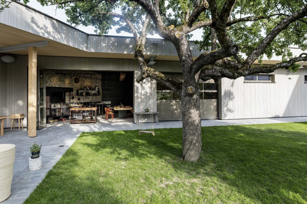 Exterior view of the Hotel Restaurant Krainer in Langenwang with vertical wooden façade ‘Wörthersee coated’ by Mareiner. A large tree with a swing stands in front of the open-plan outdoor kitchen, with a well-tended lawn and natural stone floor below.