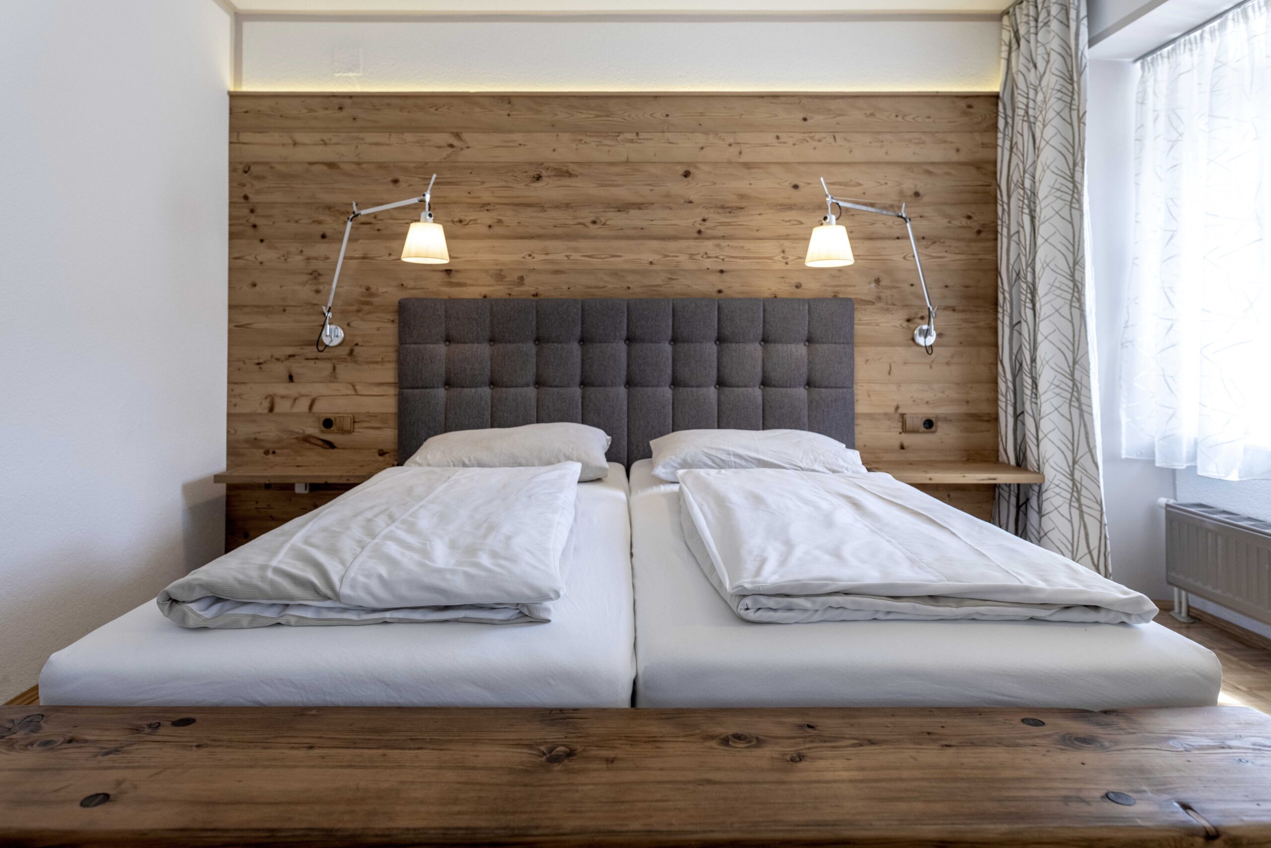 Gemütliches Doppelzimmer mit Kopfteil vor einer rustikalen Wandverkleidung aus Mareiner Gran Paradiso 212° gehackt. Die strukturierte Holzoberfläche verleiht dem Raum Wärme und Charakter. Dezente Beleuchtung und helle Stoffe sorgen für Behaglichkeit.