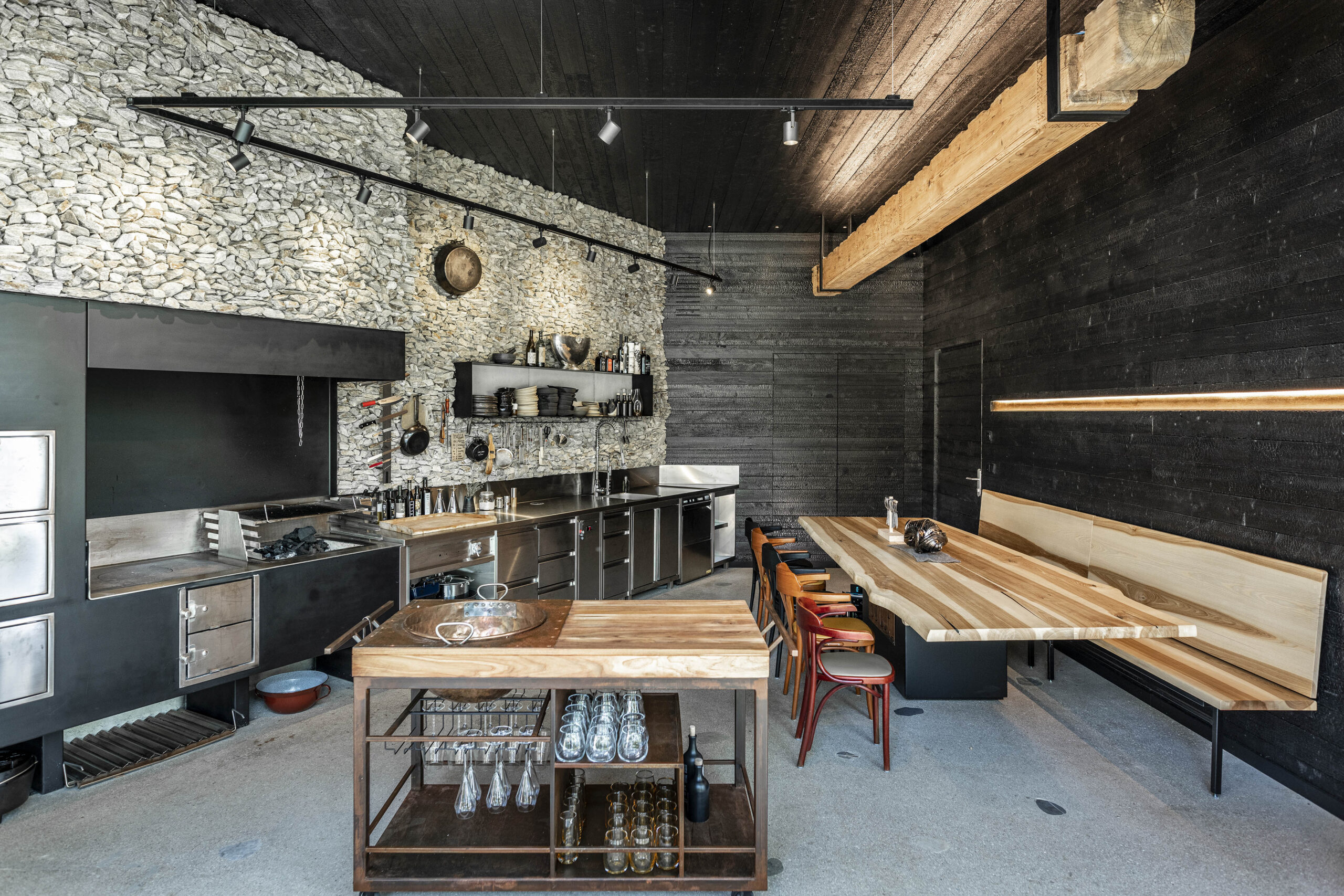 Moderne Gastroküche im Hotel Restaurant Krainer mit dunkler Vulcano-Holzverkleidung an Wänden und Decke. Natursteinwand, Edelstahlküche und große Tischplatte aus Massivholz schaffen einen stilvollen Materialmix mit hochwertigem Charakter.