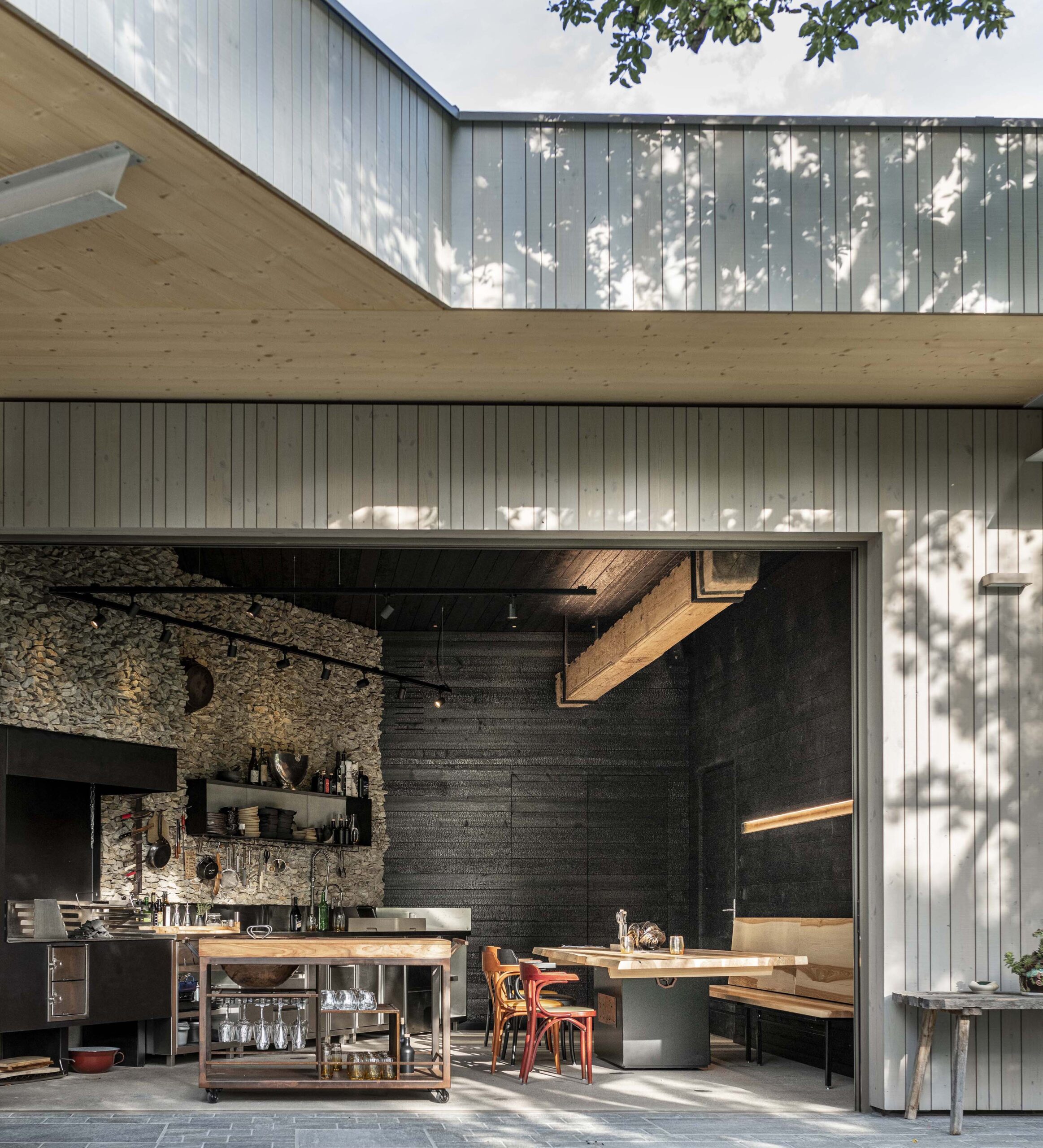 Detailansicht der offenen Küche im Hotel Restaurant Krainer: Außenfassade mit senkrechter Holzverschalung in „Wörthersee beschichtet“, Innenwände in verkohltem „Vulcano“-Holz. Natursteinwand, Holzbalken und rustikale Einrichtung runden das Bild ab.