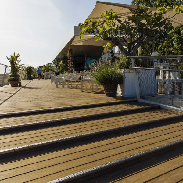 Weite Ansicht der Terrasse des Lokals „Motto am Fluss“ in Wien. Im Vordergrund breite Holzstufen mit Metallschutzkanten, darüber Kebony-Dielen von Mareiner. Weiße Möbel, üppige Bepflanzung und Sonnensegel sorgen für mediterranes Flair.
