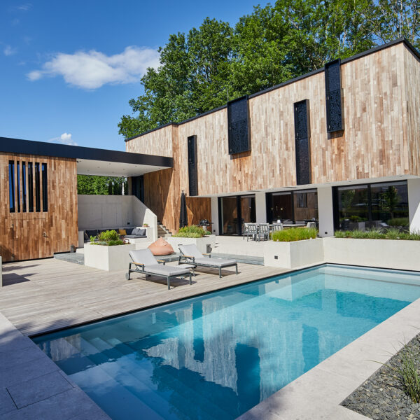 Tagesaufnahme des modernen Privathauses Blaschke mit Holzfassade von Mareiner Holz. Klare Linien, große Fensterflächen und strukturierte Ornamentverkleidungen prägen das Design. Im Vordergrund: Pool, Holzdeck, Liegen und bepflanzte Betontröge.