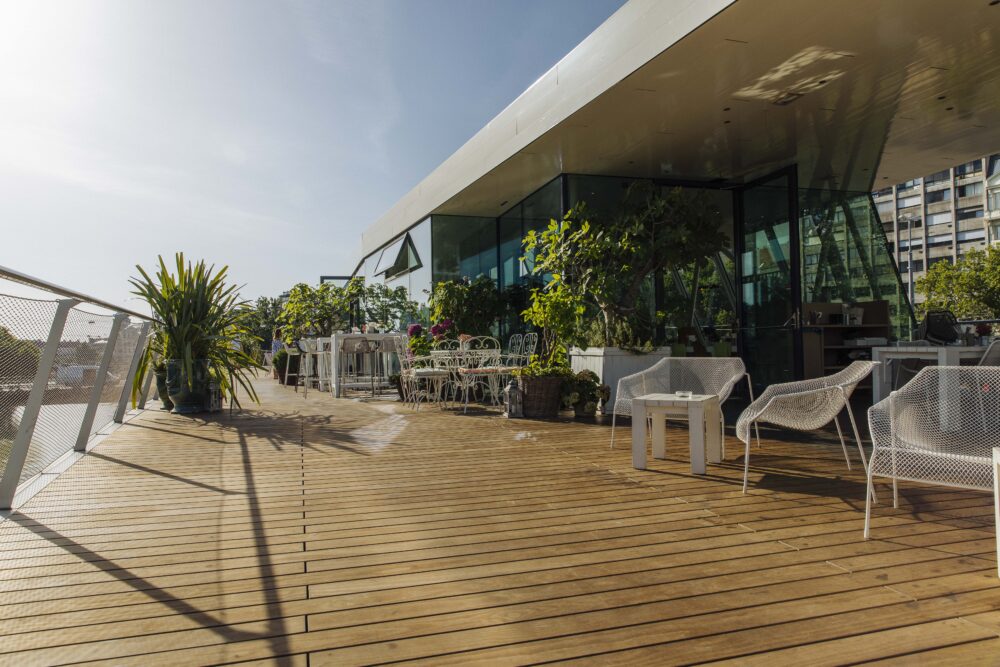 Weitläufige Terrasse des „Motto am Fluss“ in Wien mit Kebony-Holzdielen von Mareiner. Weiße Metallmöbel, zahlreiche Pflanzen und Glasfassade schaffen ein elegantes, grünes Ambiente. Die Szene ist sonnig und lädt zum Verweilen ein.