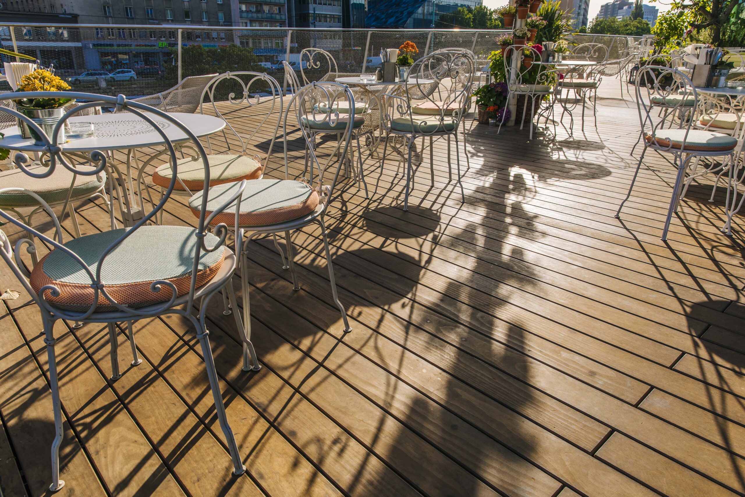 Weiße Metallstühle mit Polstern und runde Bistrotische auf der Terrasse von „Motto am Fluss“ in Wien. Der Holzboden besteht aus Kebony-Dielen von Mareiner. Lange Nachmittagsschatten und blühende Pflanzen schaffen ein romantisches Ambiente.