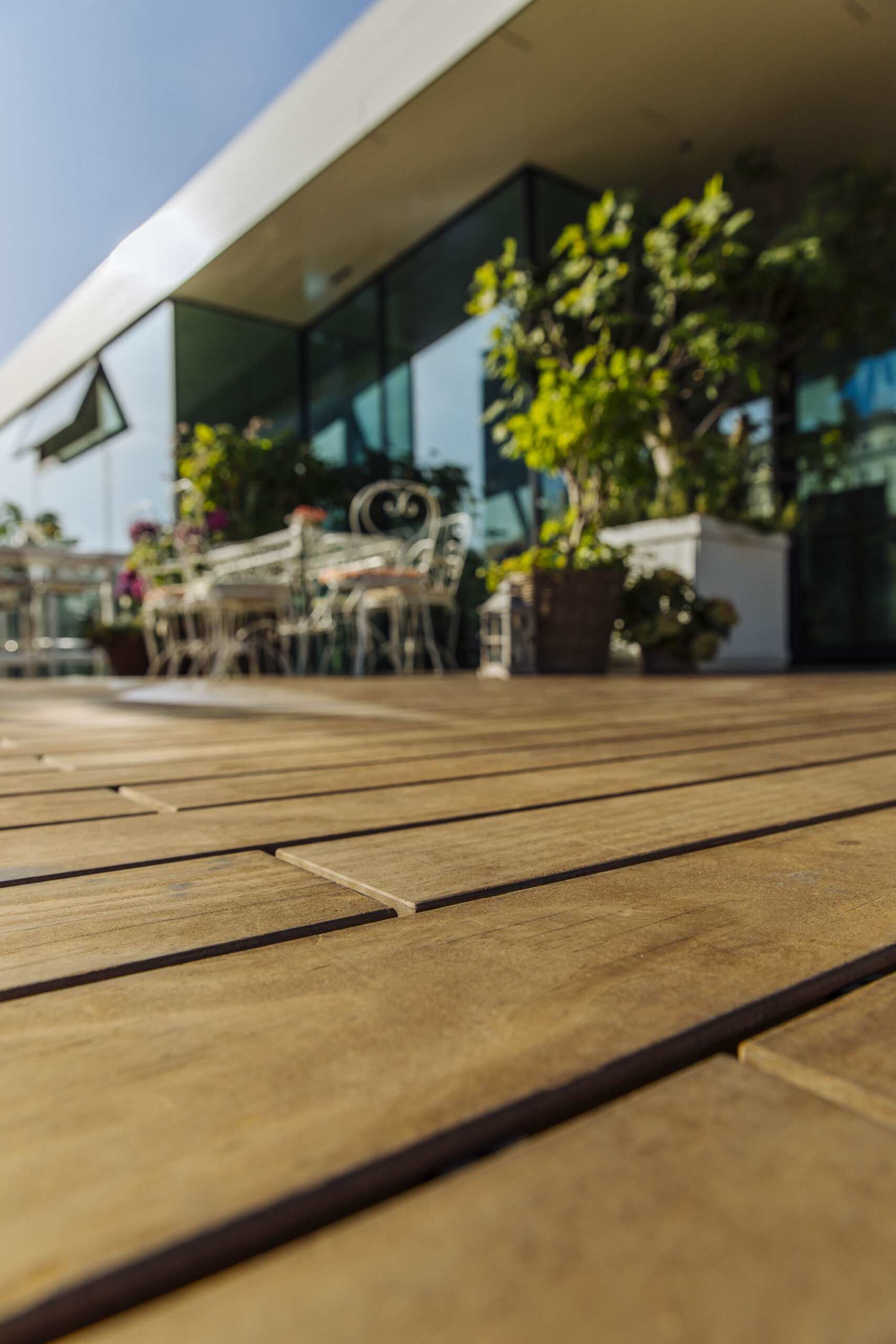 Nahaufnahme der Kebony-Holzdielen von Mareiner auf der Terrasse des Lokals „Motto am Fluss“ in Wien. Im Hintergrund unscharf: weiße Bistrostühle, Pflanzkübel und große Fensterfront mit Spiegelung. Mediterrane Atmosphäre bei Sonnenschein.
