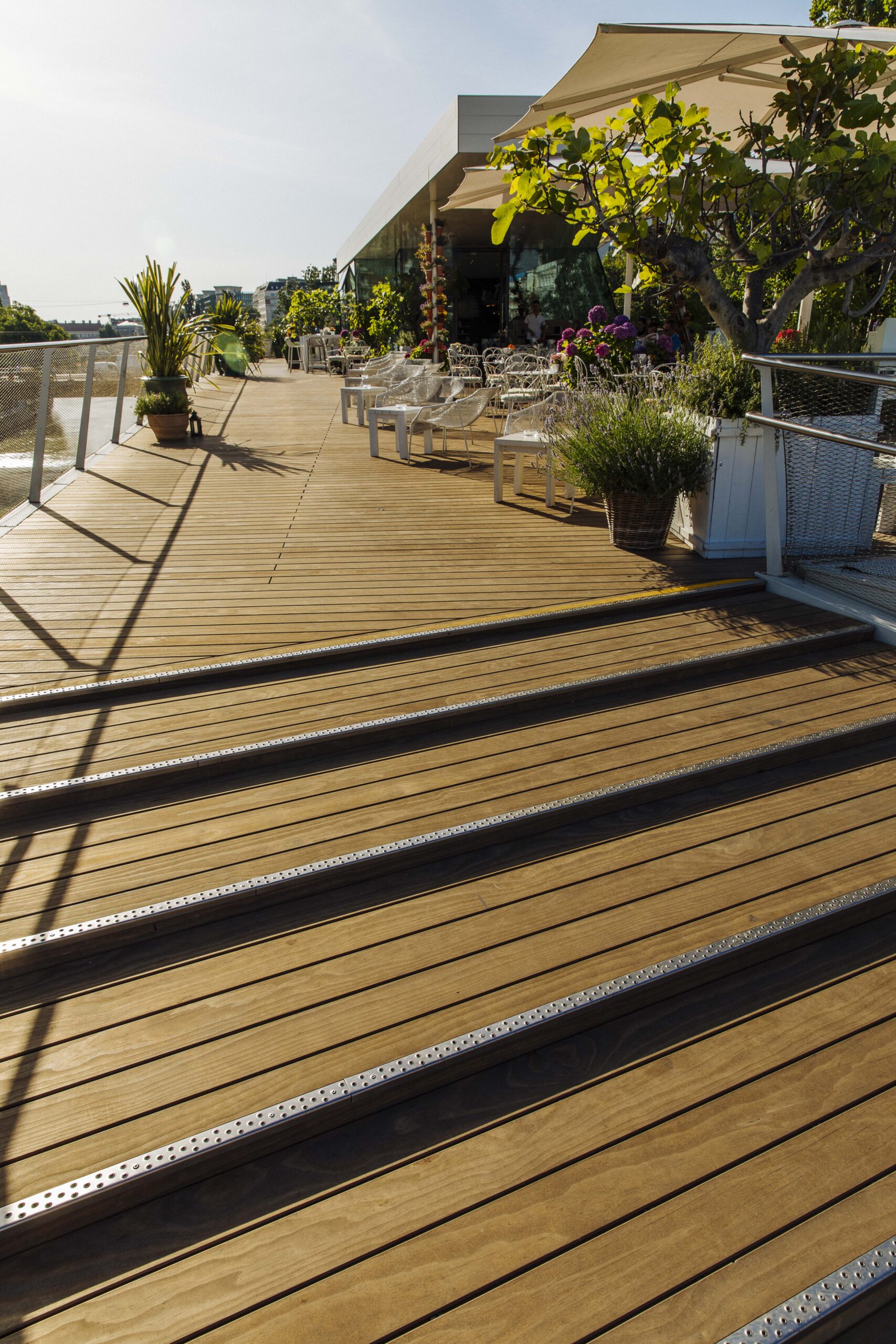 Großzügige Terrasse des Wiener Lokals „Motto am Fluss“ mit Kebony-Holzdielen von Mareiner. Im Vordergrund breite Holzstufen mit rutschfesten Metallschienen, im Hintergrund weiße Gartenmöbel, Pflanzen und Sonnensegel unter blauem Himmel.