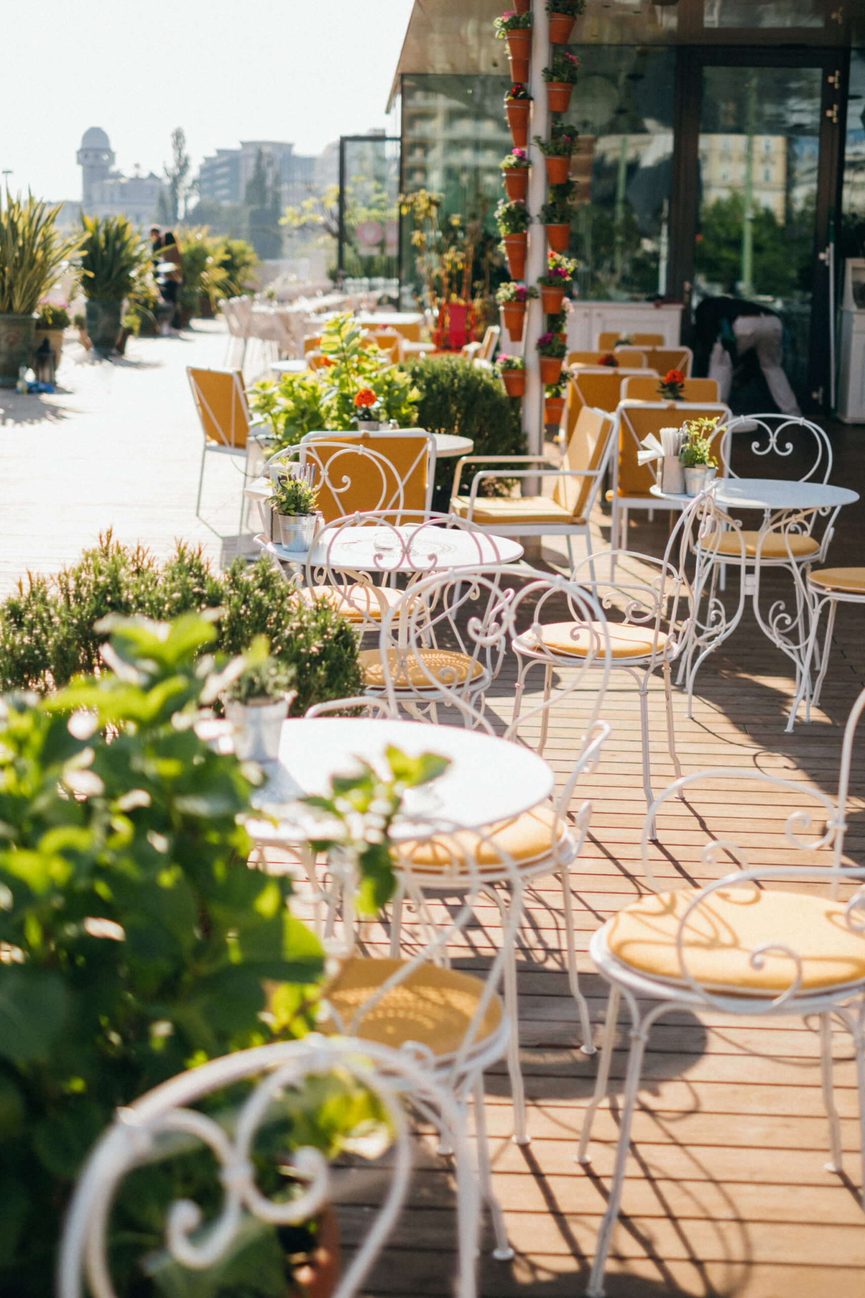 Blick auf die verspielte Terrasse des Lokals „Motto am Fluss“ in Wien. Weiße Bistrostühle mit gelben Polstern und runde Tische stehen auf Kebony-Holzdielen von Mareiner. Üppige Bepflanzung und Blumentöpfe schaffen ein charmantes, mediterranes Flair.