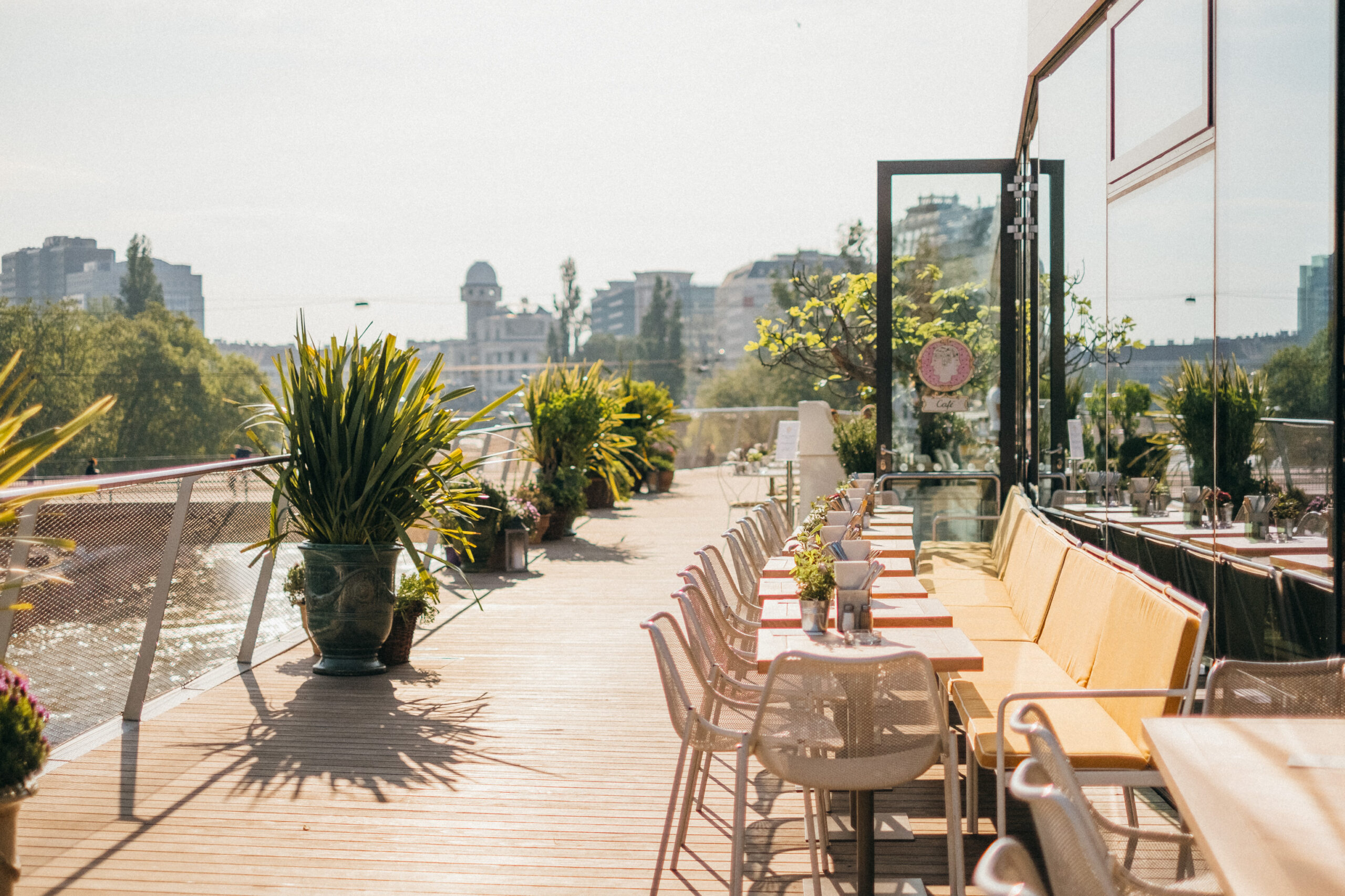 Sonnige Terrasse des Lokals „Motto am Fluss“ in Wien mit Sitzplätzen entlang der Glasfassade, gelben Polstern und Tischen. Der Boden ist mit Kebony-Holzdielen gestaltet. Üppige Pflanzkübel und Blick auf die Stadt schaffen mediterranes Flair.